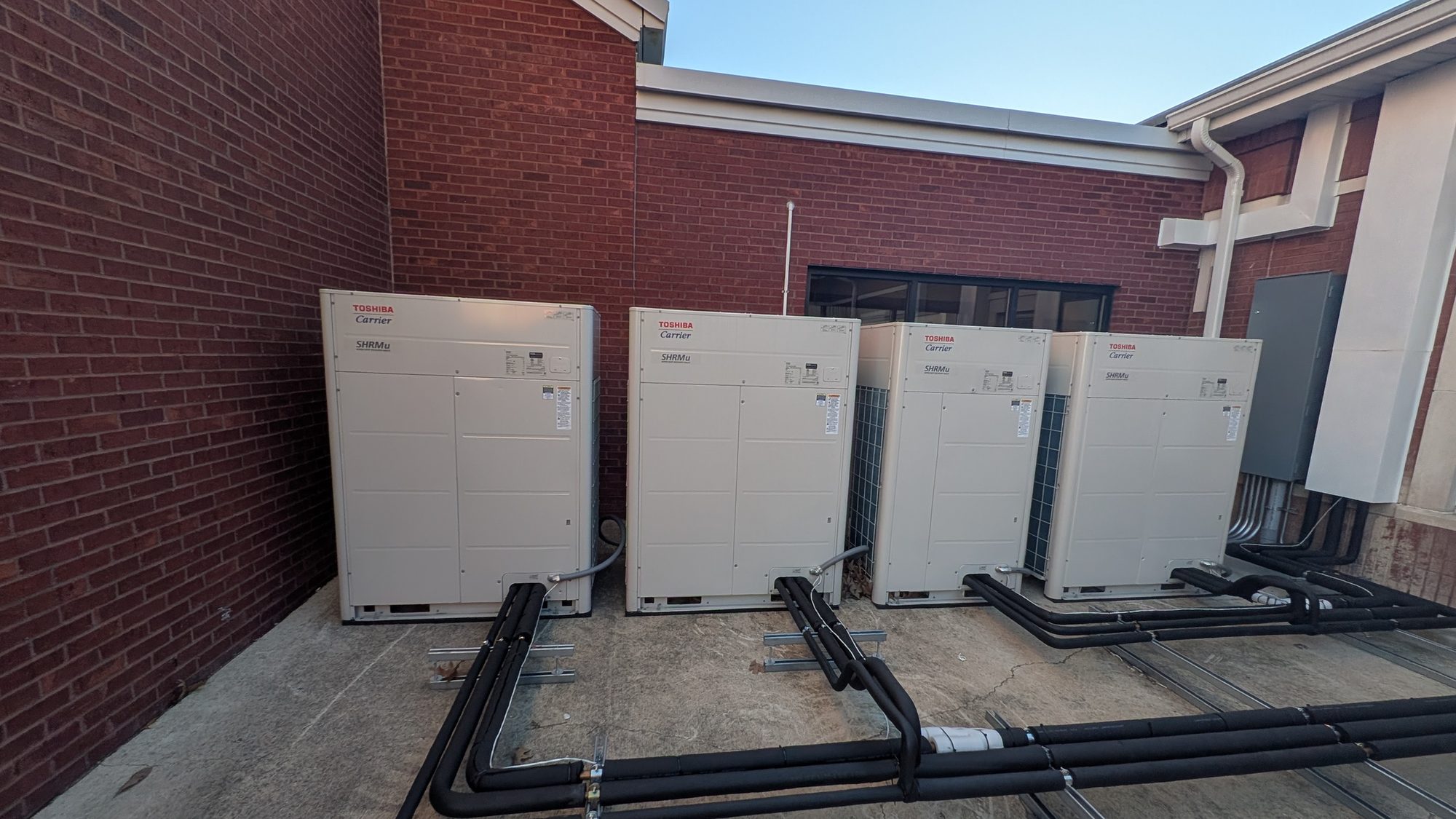 Four Toshiba-Carrier VRF condensing units on a pad beside a brick commercial building, with insulated refrigerant piping on strut